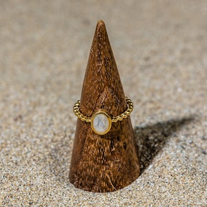 May include: A gold ring with a white oval gemstone set in a beaded band. The ring is displayed on a wooden ring holder.