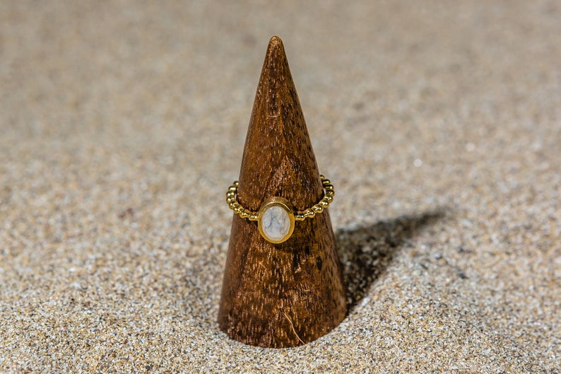 May include: A gold ring with a white oval gemstone set in a beaded band. The ring is displayed on a wooden ring holder.