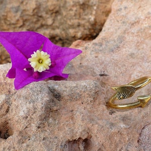 Puede incluir: Un anillo de flecha dorado con un diseño texturizado. El anillo está sobre una roca con una flor rosa.