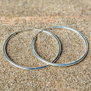 Puede incluir: Un par de pendientes de aro de plata sobre un fondo arenoso.