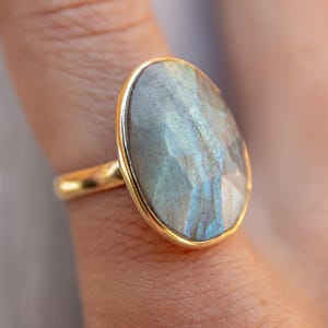 Pode incluir: Um anel de ouro com uma grande pedra oval de labradorita. A pedra tem uma cor azul, verde e cinza com flashes iridescentes.