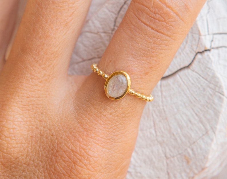 May include: A gold ring with a clear oval gemstone set in a bezel setting. The ring is made of small gold beads.