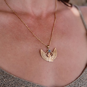 May include: Gold-toned necklace featuring a pendant with an Egyptian-inspired design. The pendant depicts a figure with outstretched wings and a small blue gemstone. The necklace is worn on a person.