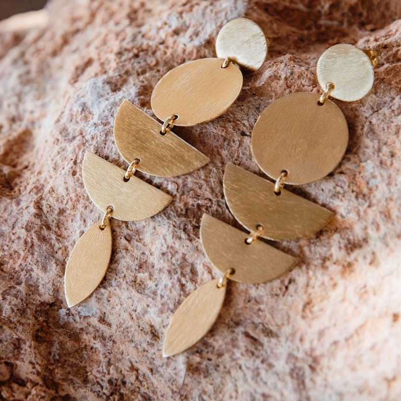 May include: A pair of gold-colored earrings with a layered design. Each earring features a small circle at the top, followed by an oval, a half-circle, and a teardrop shape. The earrings are displayed on a textured, brown surface.