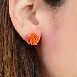 Può includere: Un paio di orecchini a bottone a forma di rosa arancione. Gli orecchini sono indossati nell'orecchio di una persona.