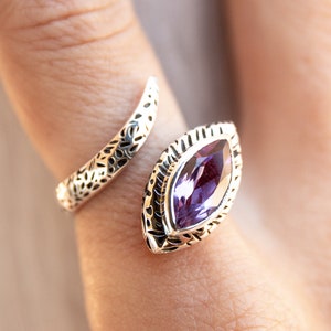May include: A silver ring with an intricate design featuring a large, purple gemstone. The ring is open and wraps around the finger.