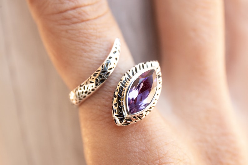 May include: A silver ring with an intricate design featuring a large, purple gemstone. The ring is open and wraps around the finger.