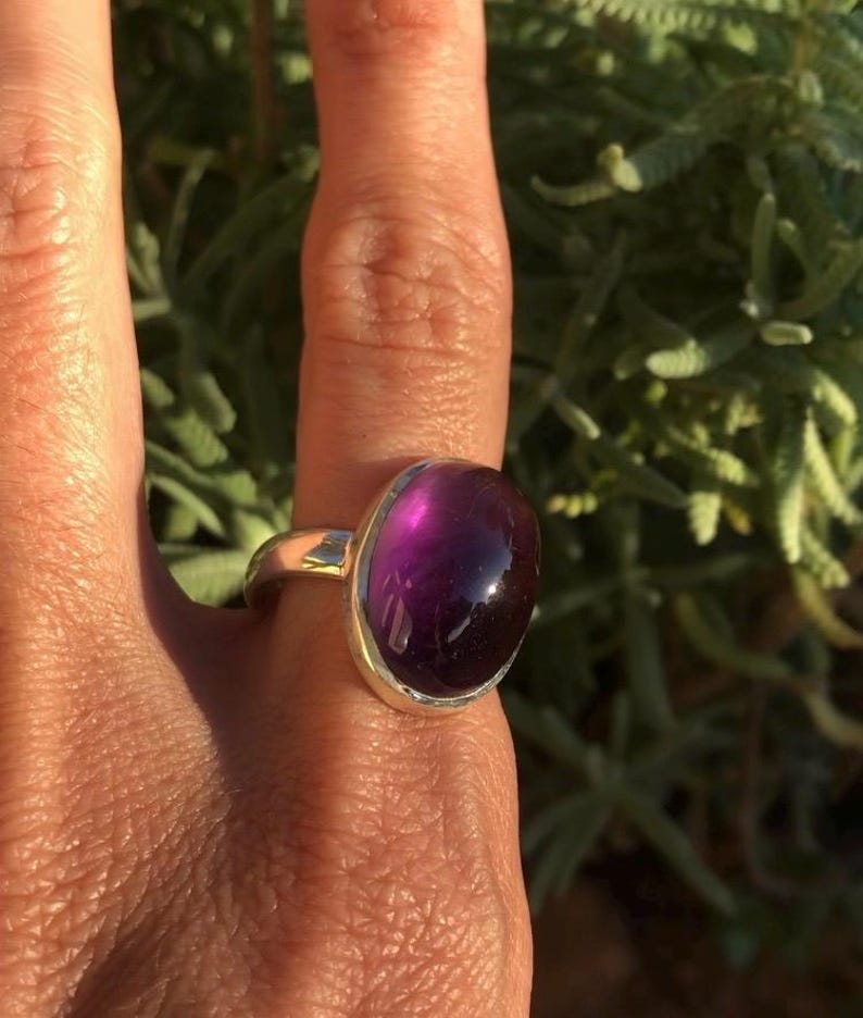 K&ouml;nnte beinhalten: Ein Silberring mit einem gro&szlig;en, ovalen, tiefvioletten Edelstein. Der polierte Edelstein hat eine gl&auml;nzende Oberfl&auml;che. Der Ring ist vor gr&uuml;nem Laub platziert, wobei Sonnenlicht die Merkmale des Rings hervorhebt.