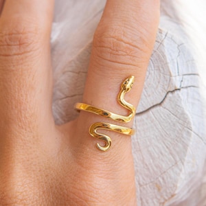 May include: A gold-coloured snake ring encircles a finger. The ring has a detailed snake head and tail, with the snake's body forming the ring's band. The ring is made of a shiny, metallic material.