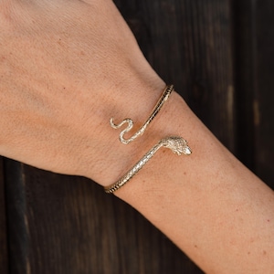 May include: A gold-toned metal cuff bracelet shaped like a coiled snake. The bracelet features a textured, scale-like pattern along the entire length of the body, with a detailed snake head at one end and a curved, tapered tail at the other end.