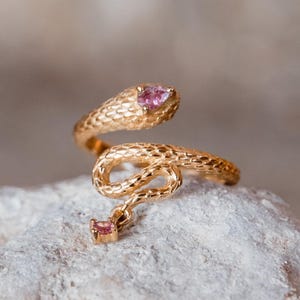 May include: A gold-colored snake ring with a textured surface. The snake's head and tail feature pink gemstones. The ring is coiled and rests on a light-colored stone surface. Jewelry.