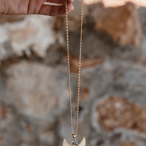 May include: Gold-colored necklace featuring a pendant with an Egyptian winged figure design. The pendant has a small blue gemstone. The necklace is suspended from a delicate chain, and is displayed against a blurred stone background.