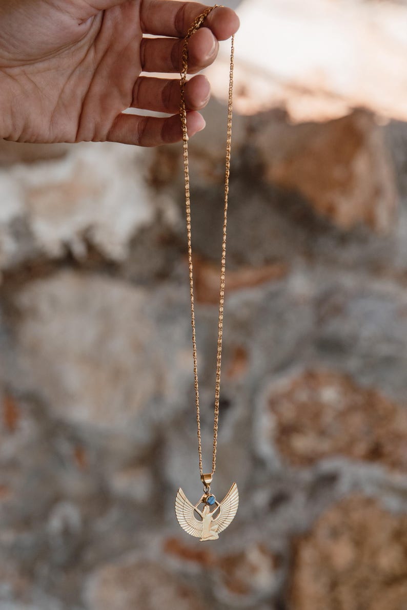 May include: Gold-colored necklace featuring a pendant with an Egyptian winged figure design. The pendant has a small blue gemstone. The necklace is suspended from a delicate chain, and is displayed against a blurred stone background.