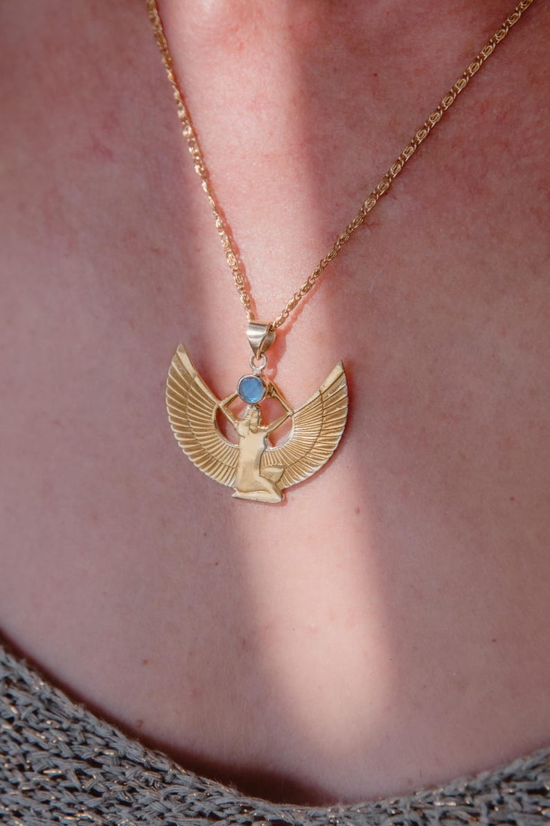 May include: Gold-colored pendant necklace featuring an Egyptian goddess design with outstretched wings and a blue gemstone. The pendant hangs from a delicate chain, creating an elegant accessory.