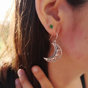 May include: Silver crescent moon earrings with intricate floral and geometric cut-out designs. The earrings hang from a simple hook and are worn on the ear. A small green stud earring is also visible.