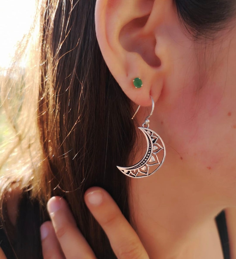 May include: Silver crescent moon earrings with intricate floral and geometric cut-out designs. The earrings hang from a simple hook and are worn on the ear. A small green stud earring is also visible.