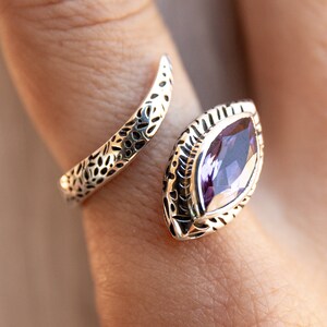 May include: A silver ring with an intricate design featuring a large, purple gemstone. The ring is open and wraps around the finger.