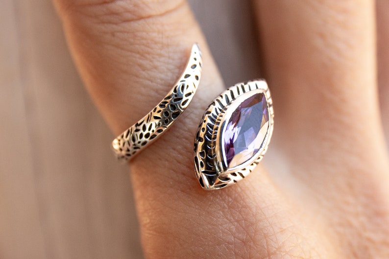 May include: A silver ring with an intricate design featuring a large, purple gemstone. The ring is open and wraps around the finger.