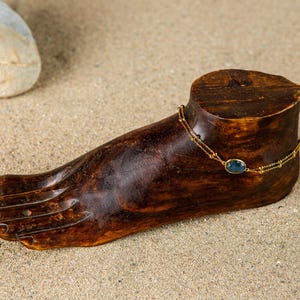 Könnte beinhalten: Ein goldener Fußkettchen mit einem blauen Edelstein wird auf einem Holzfuß ausgestellt. Das Fußkettchen besteht aus zarten Goldketten und hat einen kleinen, oval geformten blauen Stein. Der Fuß ist dunkelbraun und liegt auf einer sandigen Oberfläche.
