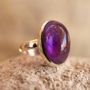 K&ouml;nnte beinhalten: Ein Silberring mit einem gro&szlig;en, oval geformten, tiefvioletten Edelstein. Der Edelstein ist in eine silberne L&uuml;nette gefasst und liegt auf einer strukturierten Oberfl&auml;che. Der Ring selbst ist ebenfalls aus Silber, was einen klassischen und eleganten Look erzeugt.