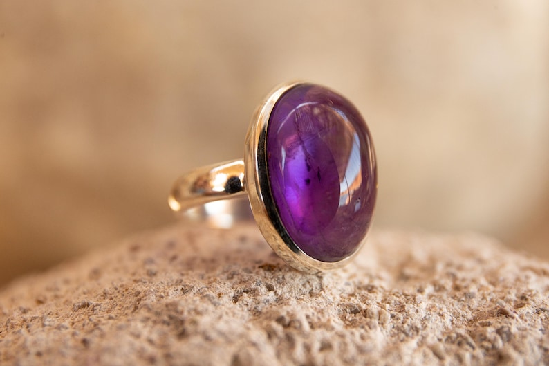 K&ouml;nnte beinhalten: Ein Silberring mit einem gro&szlig;en, oval geformten, tiefvioletten Edelstein. Der Edelstein ist in eine silberne L&uuml;nette gefasst und liegt auf einer strukturierten Oberfl&auml;che. Der Ring selbst ist ebenfalls aus Silber, was einen klassischen und eleganten Look erzeugt.