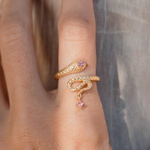 Pode incluir: Um anel de cobra dourado com uma superfície texturizada. A cabeça da cobra apresenta uma pedra rosa, e uma pequena pedra rosa pende da cauda da cobra. O anel está enrolado em volta de um dedo.
