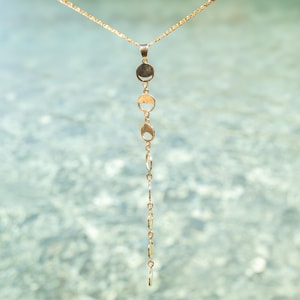 May include: A gold chain necklace with a series of small, round, gold pendants hanging from it. The necklace is photographed against a blurred background of water.