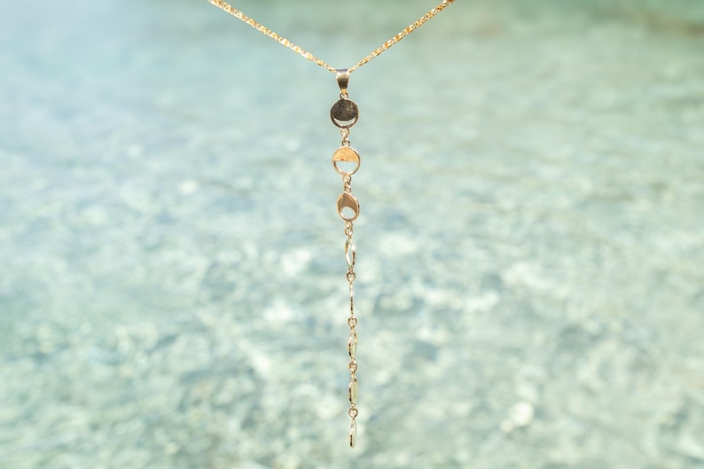 May include: A gold chain necklace with a series of small, round, gold pendants hanging from it. The necklace is photographed against a blurred background of water.