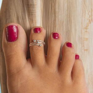 Puede incluir: Un anillo de plata para el dedo gordo del pie con tres pequeñas cuentas redondas se usa en un pie con uñas de los pies magenta pulidas. El pie descansa sobre una superficie de madera clara texturizada.