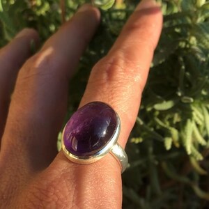 K&ouml;nnte beinhalten: Ein Silberring mit einem gro&szlig;en, ovalen Amethyst-Edelstein. Der tiefviolette Stein ist in einem polierten Silberband gefasst. Der Ring wird vor einem Hintergrund aus gr&uuml;nem Laubwerk gezeigt.