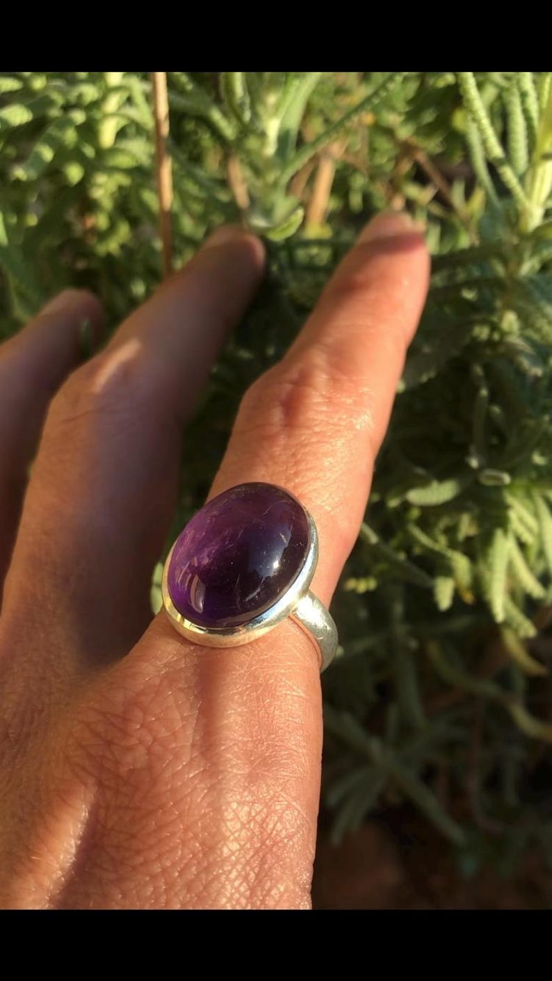 K&ouml;nnte beinhalten: Ein Silberring mit einem gro&szlig;en, ovalen Amethyst-Edelstein. Der tiefviolette Stein ist in einem polierten Silberband gefasst. Der Ring wird vor einem Hintergrund aus gr&uuml;nem Laubwerk gezeigt.