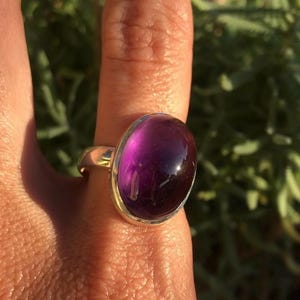 K&ouml;nnte beinhalten: Ein goldfarbener Ring mit einem gro&szlig;en, ovalen, tiefvioletten Edelstein. Der Edelstein hat eine polierte, gl&auml;nzende Oberfl&auml;che. Der Ring befindet sich an einem Finger, mit einem verschwommenen gr&uuml;nen Hintergrund.