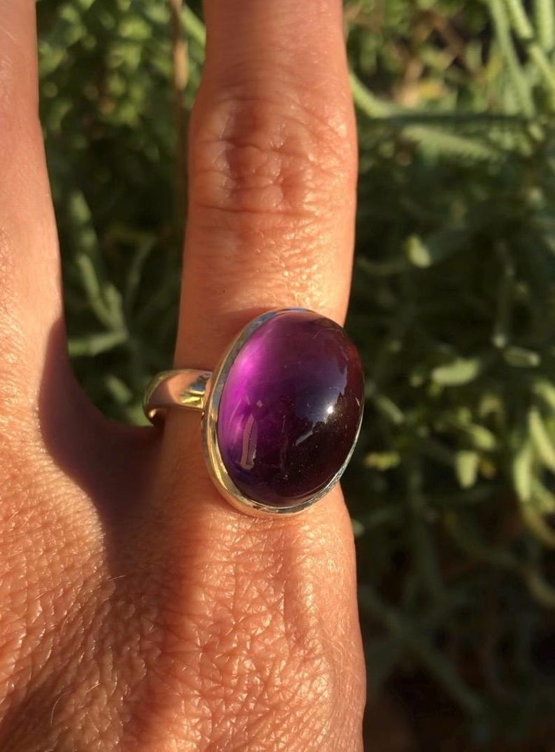 K&ouml;nnte beinhalten: Ein goldfarbener Ring mit einem gro&szlig;en, ovalen, tiefvioletten Edelstein. Der Edelstein hat eine polierte, gl&auml;nzende Oberfl&auml;che. Der Ring befindet sich an einem Finger, mit einem verschwommenen gr&uuml;nen Hintergrund.