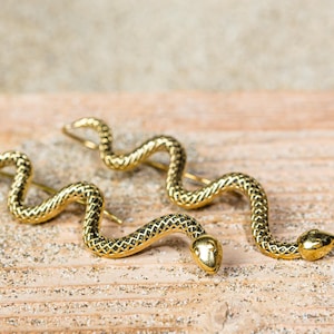 May include: A pair of gold-coloured snake earrings. The earrings are shaped like a snake, with a textured surface and a curved design. The earrings are displayed on a wooden surface with sand.