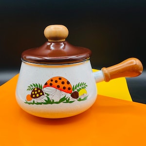 Puede incluir: Cacerola de esmalte blanco con tapa marrón y asa de madera. La cacerola presenta un diseño caprichoso de setas en naranja, marrón y amarillo. El asa y el pomo de la tapa son de madera clara. La cacerola está diseñada para cocinar.