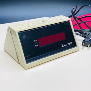 May include: A vintage beige and black digital alarm clock with a red LED display. The clock has buttons for fast, slow, time, and alarm settings. The brand name "LLOYD'S" is visible on the front. A black power cord and plug are attached.