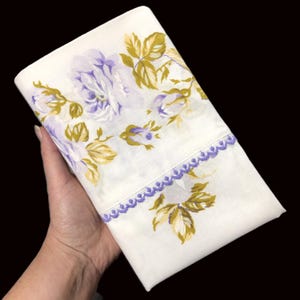 May include: White floral handkerchief with a lavender and olive green rose pattern. The handkerchief has a lavender lace trim along one edge. The fabric is folded and held in a hand.