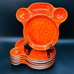Könnte beinhalten: Ein Satz von vier orangefarbenen Keramiktellern mit einem einzigartigen Design. Die Teller haben einen erhöhten Rand und eine kleine Vertiefung in der Mitte. Die Teller sind übereinander gestapelt.