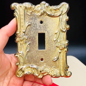 May include: Ornate gold-colored light switch cover with a rectangular opening for the switch and two screw holes. The cover features a detailed, raised floral and scroll design around the edges.
