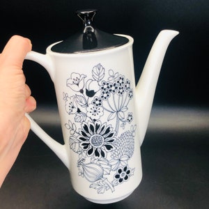 May include: White ceramic coffee pot with a black lid and a black and white floral design.