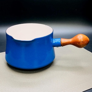 Puede incluir: Una cacerola de esmalte azul con interior blanco y asa de madera. La cacerola tiene un cuerpo redondeado y un pico vertedor. El asa es de madera marrón claro y está sujeta al lateral de la cacerola.