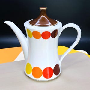 Puede incluir: Una cafetera de cerámica blanca con tapa y asa marrones. La cafetera está decorada con una serie de lunares de colores amarillo, naranja, rojo y marrón. El pico es largo y delgado.