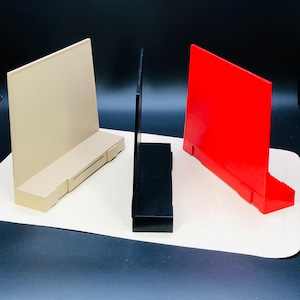 May include: Three display stands in beige, black, and red. Each stand has a rectangular base and a vertical panel. The beige stand has a shelf-like feature. The stands are arranged on a white surface against a dark background.