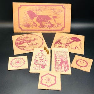 Puede incluir: Un conjunto de nueve placas de madera con varias imágenes impresas en rosa. Las imágenes incluyen un perro, dos caballos, un gato y un perro, un payaso, un indígena y tres diseños florales diferentes. También hay cinco tachuelas pequeñas de latón.