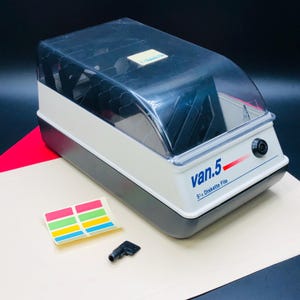 Könnte beinhalten: Ein Vintage-Diskettenarchiv in Grau, Weiß und Schwarz für 5 1/4" Disketten mit einem Deckel aus transparentem Kunststoff. Das Archiv trägt den Text "van.5" und enthält einen Aufkleberbogen und einen kleinen schwarzen Schlüssel.