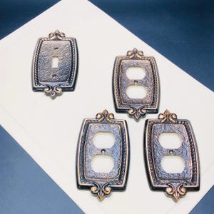 Könnte beinhalten: Vier dekorative Steckdosenabdeckungen in Bronze mit einem verzierten Design. Die Abdeckungen haben eine strukturierte Oberfläche und ein Fleur-de-lis-Design oben.