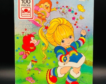 Puede incluir: Una caja de rompecabezas vintage de 100 piezas con una colorida ilustración de un personaje de dibujos animados con cabello rubio, un vestido azul y detalles de arcoíris. La caja tiene el logotipo Golden y las palabras "Jigsaw Puzzle" y "Casse-Tete".