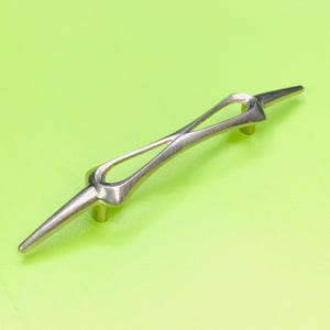 May include: A silver-coloured cabinet pull with a distinctive, open design. The handle has a central, elongated oval shape with pointed ends. It is mounted on two small, cylindrical posts, and is set against a bright green background.