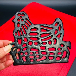 以下が含まれることがあります： 切り抜きデザインの黒い鋳鉄製鶏型トリベット。赤いまな板の上に置かれています。鶏の形をしたトリベットは、熱い調理器具から表面を保護するように設計されています。