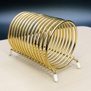 May include: A gold-colored metal napkin holder with a circular design. The holder has a rectangular base with four white-tipped feet. The circular section is made of multiple curved metal rods.